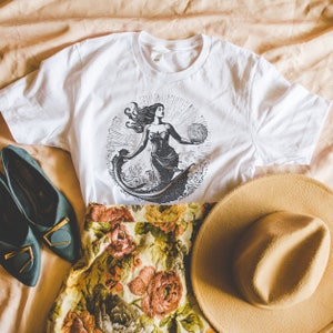 May include: White t-shirt with a black and white illustration of a mermaid holding a globe. The mermaid is surrounded by waves. The t-shirt is laying on a floral patterned skirt and a brown hat.