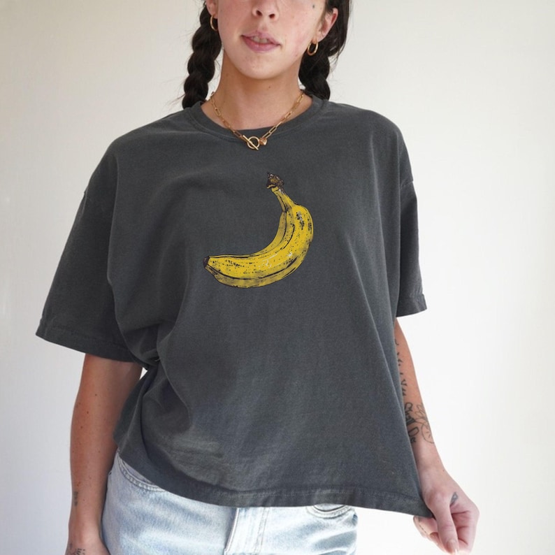 May include: Dark gray t-shirt featuring a yellow banana graphic. The banana is centrally placed on the shirt. The shirt has short sleeves and a relaxed fit. The model is wearing a gold necklace and blue jeans.