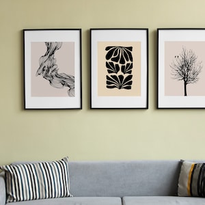 May include: Three black and white framed prints on a pale green wall above a grey sofa with striped and patterned cushions. The prints feature abstract designs, including a swirling pattern, a floral design, and a bare tree.
