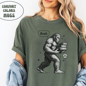 Può includere: T-shirt verde muschio con grafica di un Bigfoot che trasporta libri. Il Bigfoot ha un'espressione seria e tiene tre libri. Il testo "Book." è sopra la testa del Bigfoot. La maglietta ha anche il testo "the collector".