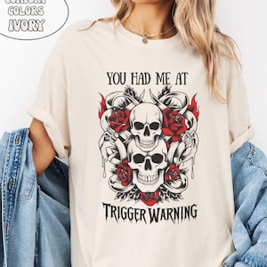 May include: Ivory-colored t-shirt featuring a graphic of two skulls, red roses, and the text "YOU HAD ME AT TRIGGER WARNING." The design includes flames and decorative elements, creating a gothic aesthetic.