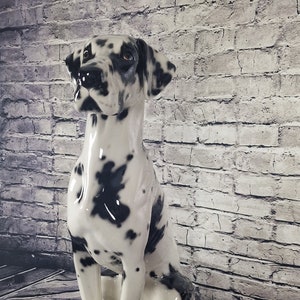 May include: A ceramic Dalmatian dog figurine in a seated position. The dog has a glossy white coat with black spots. The figurine has a detailed face and is set against a brick wall and wooden floor backdrop.