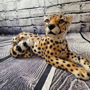 Könnte beinhalten: Eine Keramikfigur eines Geparden mit einem gefleckten braunen und schwarzen Muster. Der Gepard liegt mit dem Kopf zur Seite gedreht und den Pfoten unter dem Körper.