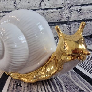 May include: A white ceramic snail sculpture with a gold-painted body and shell. The shell has a detailed spiral pattern.