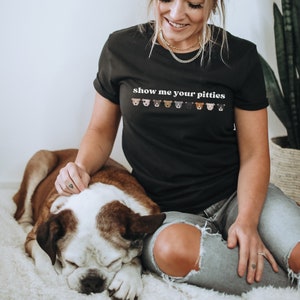 Pode incluir: Uma mulher vestindo uma camiseta preta com o texto "show me your pitties" e uma fileira de rostos de pit bull impressos abaixo. Ela está sentada em um tapete branco e felpudo com um cachorro marrom e branco deitado ao lado dela.