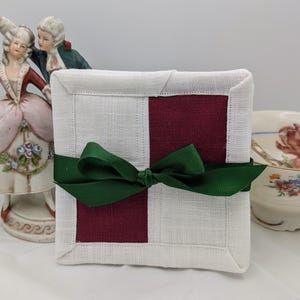 Könnte beinhalten: Quadratische Untersetzer in Weiß und Burgunderrot, gebunden mit einem dunkelgrünen Band. Die Untersetzer haben einen genähten Rand und ein zweifarbiges Design. Der Hintergrund umfasst Porzellanfiguren und eine Blumenvase.