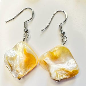 May include: A pair of drop earrings featuring irregular, diamond-shaped pieces of iridescent shell with swirling white and golden-yellow tones. The shell pendants are suspended from silver-toned metal fishhook wires with small coiled accents.