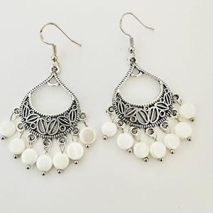 May include: A pair of silver-toned dangle earrings featuring a teardrop-shaped frame with intricate cut-out leaf patterns. Each earring is adorned with multiple small, round, white shell-like discs suspended from the bottom edge on silver hooks.