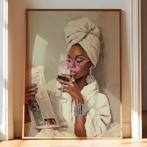 May include: A framed art print shows a woman in a white robe and towel, wearing pink sunglasses and large earrings, holding a glass of red wine and reading a newspaper. The artwork has a gold frame and is leaning against a wall.
