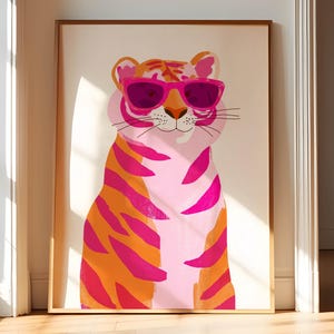 May include: A framed art print of a tiger wearing pink sunglasses. The tiger is depicted with pink and orange stripes on a light pink body. The artwork is displayed in a room with a wooden floor and a white wall.