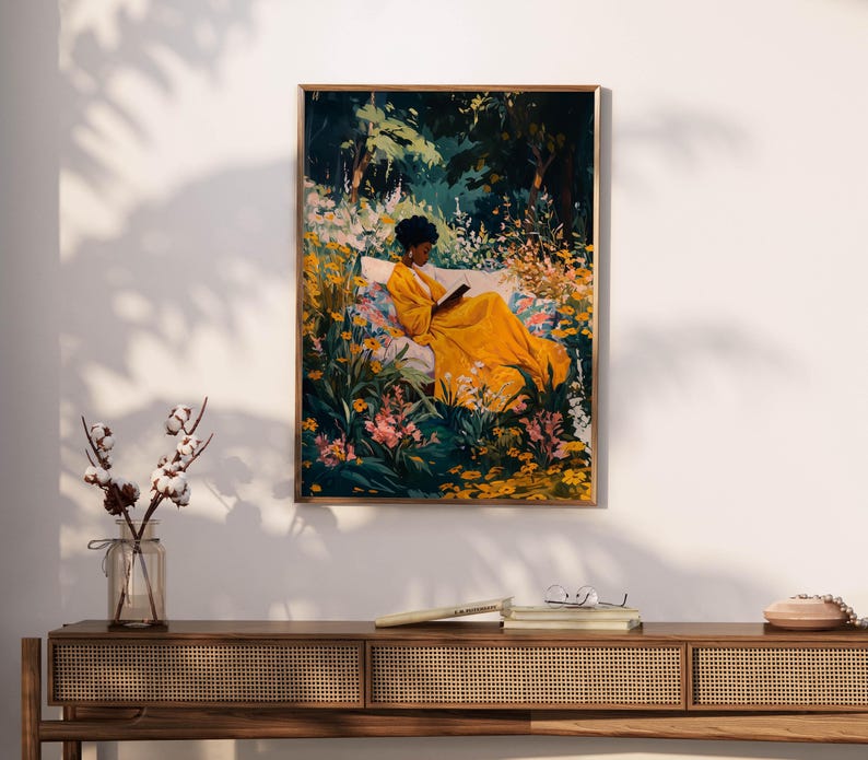 May include: A framed art print depicts a person in a yellow dress reading in a garden. The artwork features a person sitting on a bench surrounded by colorful flowers and lush greenery. The print is displayed on a white wall above a wooden console.