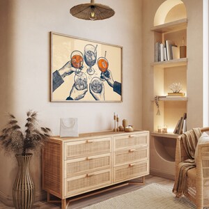 Puede incluir: Una ilustraci&oacute;n enmarcada de cuatro manos sosteniendo vasos de bebidas, con un fondo beige claro. La ilustraci&oacute;n est&aacute; colgada en una pared sobre una c&oacute;moda de madera clara con seis cajones.