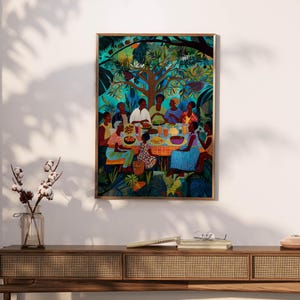 May include: A framed art print showing a colourful outdoor gathering. People are seated around a table with food, under a large tree. The artwork uses a vibrant palette of blues, greens, and oranges, creating a lively scene. The print is in a wooden frame.