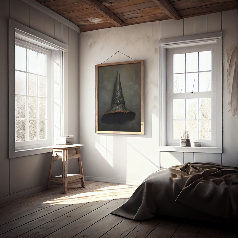 Op de afbeelding: Een slaapkamer met een houten vloer, witte muren en twee ramen. Een bed met een bruin dekbed staat op de voorgrond. Een ingelijst schilderij van een zwarte heksenhoed hangt aan de muur. Een kleine houten kruk met boeken erop staat in de hoek van de kamer.