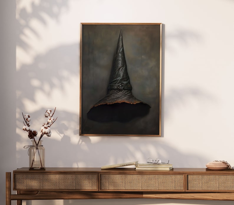 Op de afbeelding: Een ingelijst schilderij van een zwarte heksenhoed met een puntige top en een brede rand. De hoed is geplaatst tegen een donkergrijze achtergrond met subtiele hints van lichtblauw.