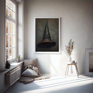 Op de afbeelding: Een ingelijst schilderij van een zwarte heksenhoed die aan een muur hangt in een kamer met een raam, een bank en een bijzettafel met een vaas met gedroogde bloemen.
