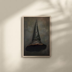 Op de afbeelding: Een ingelijst schilderij van een zwarte, puntige heksenhoed met een bruine rand tegen een donkergrijze achtergrond.