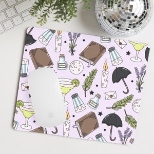 May include: A purple mouse pad with a repeating pattern of black umbrellas, books, candles, salt shakers, potion bottles, sprigs of lavender, and cocktails.