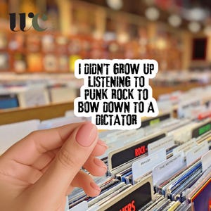 May include: A white sticker with black text that reads "I DIDN'T GROW UP LISTENING TO PUNK ROCK TO BOW DOWN TO A DICTATOR." The sticker is held in front of a collection of vinyl records.