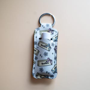 May include: A white lip balm holder with a silver key ring. The holder has a repeating pattern of butter sticks, flowers, and bows. A white lip balm is visible in the holder's opening. The background is a solid tan color.