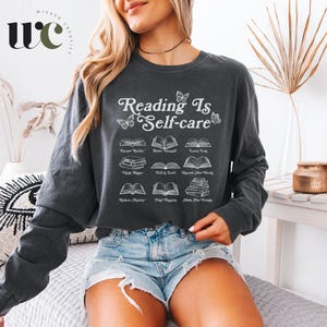 Op de afbeelding: Donkergrijs shirt met lange mouwen met de tekst "Reading is Self-care" en illustraties van boeken. Het shirt is gemaakt van een zacht materiaal en is geschikt voor vrijetijdskleding. Het shirt heeft een comfortabele pasvorm.