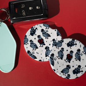 May include: A black car key fob, a light mint green motel-style keychain, and two round white car coasters featuring a repeating pattern of cartoon pigeons with mohawks, guitars, and red hearts, all set against a solid red background.