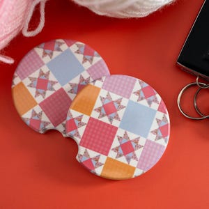 May include: Two circular car coasters featuring a repeating quilt-inspired pattern of pink, light blue, and orange squares interspersed with multi-colored star motifs. Each coaster has a small semi-circular notch along the edge for easy removal.