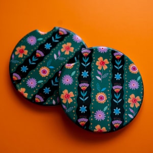 May include: Two floral car coasters. The coasters are round with a dark green background and a pattern of colorful flowers, stars, and black stripes. The coasters are on an orange background.