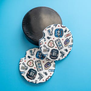 May include: Two white car coasters with a hockey theme, featuring a black hockey puck on a blue background. The coasters have a repeating pattern of hockey jerseys, phones, and other hockey-related graphics.