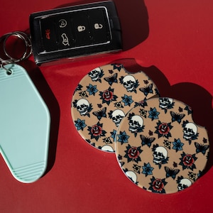 May include: A light blue motel-style keychain, a black car key fob, and two circular car coasters featuring a tan background with a repeating pattern of white skulls, red roses, blue flowers, and black butterflies on a red surface.