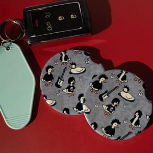 May include: A light mint green motel-style keychain, a black car key fob, and two circular car coasters featuring a repeating pattern of cartoon ducks with punk rock hairstyles, electric guitars, and skull shirts on a gray background.