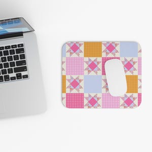 May include: A colorful mousepad with a patchwork quilt design. The pattern features squares of pink, orange, and blue, with floral star accents. A white computer mouse rests on the pad, next to a laptop.
