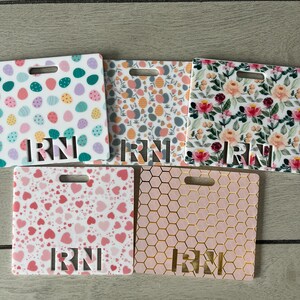 Peut inclure: Cinq étiquettes de nom carrées à motifs différents avec les lettres "RN" découpées. Les motifs comprennent des œufs de Pâques pastel, des fleurs, des cœurs et un motif en nid d'abeille.