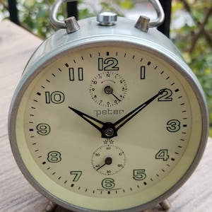 Könnte beinhalten: Ein silberner Wecker mit weißem Zifferblatt und schwarzen Zeigern. Die Uhr hat einen kleinen Sekundenzeiger und einen größeren Minutenzeiger. Das Zifferblatt der Uhr hat die Zahlen 1 bis 12 und das Wort "peter" darauf gedruckt. Die Uhr ist in Deutschland hergestellt.