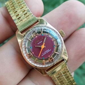 May include: A vintage gold-plated VIALUX Super watch with a burgundy face and gold hands. The watch has a clear, textured crystal and a gold link strap. The watch face displays the text "17 Jewels Super-Shock".
