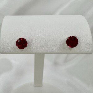 May include: A pair of stud earrings with round, deep red gemstones set in silver-coloured metal. The earrings are displayed on a white stand against a white, textured background. The gemstones have a vibrant, polished appearance.