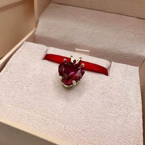 May include: A red heart-shaped pendant with a gold setting, displayed in a white box with a red ribbon.