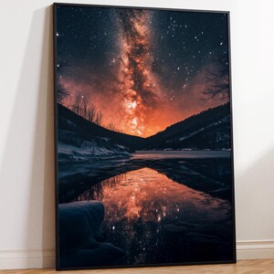 May include: A framed print featuring a nighttime landscape with a vibrant orange and red Milky Way galaxy dominating the sky. The galaxy reflects in the still water of a lake or river, with dark mountains on either side. The print is in a black frame.