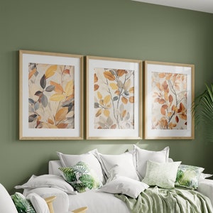 Może przedstawiać: Trzy oprawione wydruki jesiennych liści w odcieniach brązu, żółci i pomarańczy, wiszące na zielonej ścianie nad białą sofą z zielonymi poduszkami i zielonym kocem.