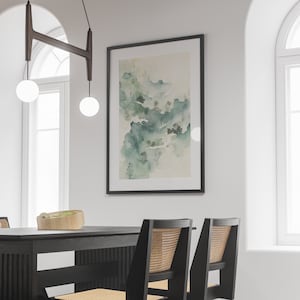May include: A black framed print of a watercolor painting depicting a landscape with green and white hues. The print is hanging on a white wall in a dining room with a black wooden table and chairs.