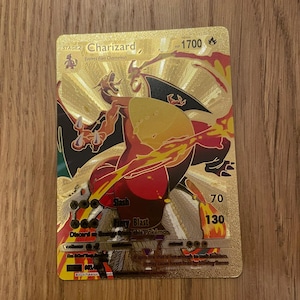 May include: A gold Pokemon trading card featuring Charizard, a fire-type Pokemon with orange and red flames. The card has a HP of 1700 and shows the attacks Slash and Fiery Blast. The text on the card reads "It spits fire that is hot enough to melt boulders. It may cause forest fires by blowing flames."