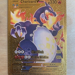 May include: Gold Charizard VMAX Pokemon card featuring a black and purple illustration of Charizard. The card has a gold background and displays the text "Charizard VMAX" and "HP 330". The card also has two attacks: "Claw Slash" for 100 damage and "G-Max Wildfire" for 300 damage. The card is from the Sword & Shield expansion.