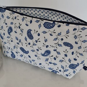 Peut inclure: Une trousse de maquillage blanche ornée d'un motif floral cachemire bleu. Elle est dotée d'une fermeture éclair noire, d'un pompon bleu et d'une petite poignée. L'intérieur est doublé d'un motif floral bleu et blanc.