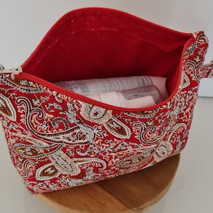 Peut inclure: Une trousse de maquillage matelassée à motif paisley rouge avec une fermeture à glissière et une poignée. La trousse est ouverte et contient plusieurs tubes et flacons blancs.