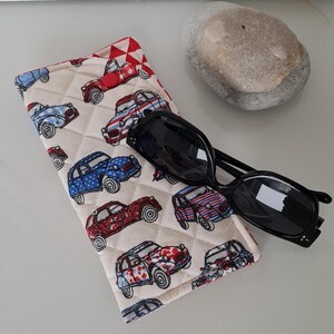 Peut inclure: Un étui à lunettes matelassé blanc avec une doublure à carreaux rouge et blanc. L'étui est décoré d'un motif de voitures anciennes bleues et rouges. Une paire de lunettes de soleil noires est posée sur l'étui.