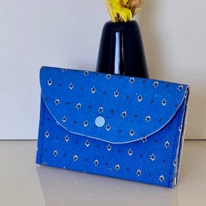 Peut inclure: Un portefeuille en tissu bleu avec une fermeture à rabat et un bouton bleu clair. Le portefeuille est orné d'un motif floral blanc. Un vase noir avec des fleurs jaunes est en arrière-plan.