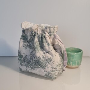 Peut inclure: Un petit sac à cordon matelassé avec un motif de toile de Jouy vert et blanc. Le sac est à côté d'une petite tasse en céramique vert clair. Le cordon du sac est de couleur violet clair.
