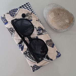 Peut inclure: Un étui à lunettes matelassé beige avec une fermeture à bouton noir. L'étui présente un motif répété de baskets colorées. Des lunettes de soleil noires sont à l'intérieur de l'étui.