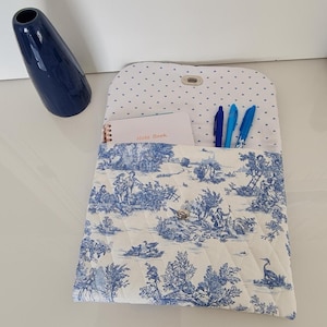 Peut inclure: Une pochette en tissu blanc avec un motif toile bleu et blanc. La pochette a un rabat qui se ferme avec un bouton. La pochette est remplie d'un carnet et de stylos.