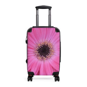 May include: A pink rolling suitcase featuring a close-up of a gerbera daisy. The suitcase has a black handle and frame, with black wheels. The flower's center is dark, surrounded by yellow and pink petals.